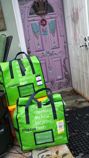 Two green Amazon Fresh delivery bags are placed on the ground in front of a uniquely painted purple door with decorative elements such as a crown and wings. The bags display various grocery items listed on them in white text. The surrounding area includes orange crates and a black mat, with the bags leaning against a wall.