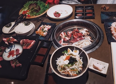 A variety of dishes are arranged on a table around an active grill. Fresh cuts of meat are cooking on the grill. There are also plates of raw and cooked foods including salad, sushi, and a vegetable platter. Small dishes of sauces are placed around the table.