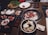A variety of dishes are arranged on a table around an active grill. Fresh cuts of meat are cooking on the grill. There are also plates of raw and cooked foods including salad, sushi, and a vegetable platter. Small dishes of sauces are placed around the table.