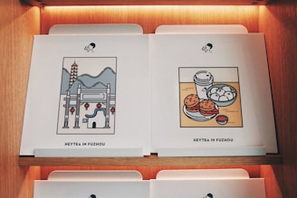 Two pieces of artwork are displayed on a wooden shelf, each featuring a minimalist style illustration. The left piece depicts a traditional Chinese architectural scene with a pagoda, mountains, and red lanterns. The right piece shows a food scene with a cup of tea, steamed buns, burgers, and a small bowl of sauce. Both have the caption 'Heytea in Fuzhou' at the bottom.