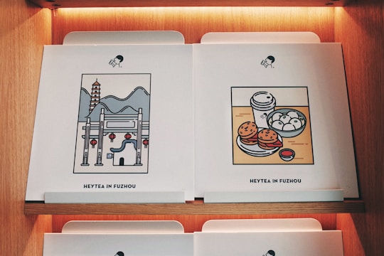 Two pieces of artwork are displayed on a wooden shelf, each featuring a minimalist style illustration. The left piece depicts a traditional Chinese architectural scene with a pagoda, mountains, and red lanterns. The right piece shows a food scene with a cup of tea, steamed buns, burgers, and a small bowl of sauce. Both have the caption 'Heytea in Fuzhou' at the bottom.
