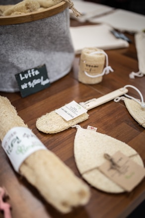 Various natural loofah products are displayed on a wooden table. These include several sponge-like items, some rolled, others cut into pads or mitts. A large container holds additional loofahs, and small labels with descriptions are attached to some products. A small chalkboard sign with text is placed in front of the container.