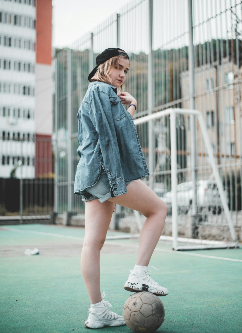 woman wearing blue denim jacket standing and stepping right foot on white soccer ball in green field during daytime