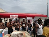 A group of muscular men standing under a red canopy, some holding bottles and papers, with promotional posters in the background. The setting appears to be an outdoor event or exhibition.
