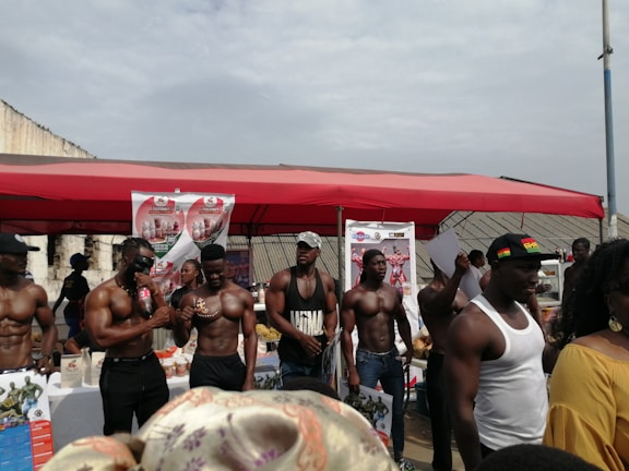 A group of muscular men standing under a red canopy, some holding bottles and papers, with promotional posters in the background. The setting appears to be an outdoor event or exhibition.