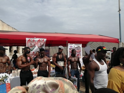 A group of muscular men standing under a red canopy, some holding bottles and papers, with promotional posters in the background. The setting appears to be an outdoor event or exhibition.