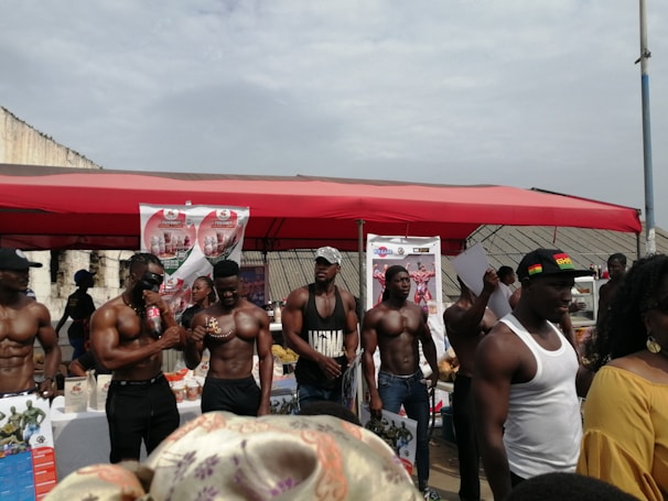 A group of muscular men standing under a red canopy, some holding bottles and papers, with promotional posters in the background. The setting appears to be an outdoor event or exhibition.