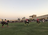 A group of polo players on horseback are actively engaged in a match on a grassy field. They wear brightly colored jerseys and helmets, with one player in blue and others in red. An ancient architectural backdrop with domes and arches surrounds the field, and spectators are gathered around watching the event.