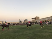 A group of polo players on horseback are actively engaged in a match on a grassy field. They wear brightly colored jerseys and helmets, with one player in blue and others in red. An ancient architectural backdrop with domes and arches surrounds the field, and spectators are gathered around watching the event.