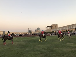 A group of polo players on horseback are actively engaged in a match on a grassy field. They wear brightly colored jerseys and helmets, with one player in blue and others in red. An ancient architectural backdrop with domes and arches surrounds the field, and spectators are gathered around watching the event.