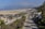 A wide expanse of sandy beach with several cars parked near the shore. A road runs parallel to the beach lined with palm trees. The background features distant mountains and a few scattered buildings under a clear, blue sky with some clouds.
