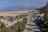 A wide expanse of sandy beach with several cars parked near the shore. A road runs parallel to the beach lined with palm trees. The background features distant mountains and a few scattered buildings under a clear, blue sky with some clouds.