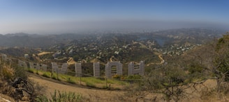 A panoramic view from behind the iconic Hollywood sign, showing a sprawling cityscape under a hazy sky. The sign's large white letters are prominently displayed along the edge of a barren hillside, with lush green vegetation in the foreground. The city of Los Angeles stretches out below, dotted with houses, winding roads, and a large body of water visible in the mid-ground.