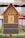 A wooden insect hotel featuring a triangular roof filled with bark and wood shavings, multiple circular wooden holes, and cylindrical bamboo tubes. The lower sections contain different natural materials and are enclosed with mesh.