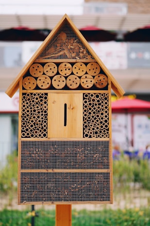 A wooden insect hotel featuring a triangular roof filled with bark and wood shavings, multiple circular wooden holes, and cylindrical bamboo tubes. The lower sections contain different natural materials and are enclosed with mesh.