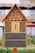 A wooden insect hotel featuring a triangular roof filled with bark and wood shavings, multiple circular wooden holes, and cylindrical bamboo tubes. The lower sections contain different natural materials and are enclosed with mesh.