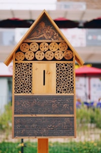 A wooden insect hotel featuring a triangular roof filled with bark and wood shavings, multiple circular wooden holes, and cylindrical bamboo tubes. The lower sections contain different natural materials and are enclosed with mesh.