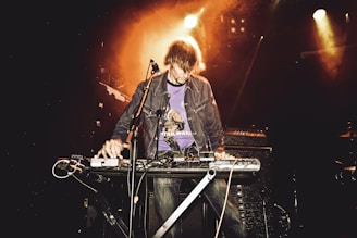 A person is performing on stage with a keyboard and various electronic equipment, surrounded by a warm, orange glowing light. They are wearing a jacket and a graphic t-shirt.