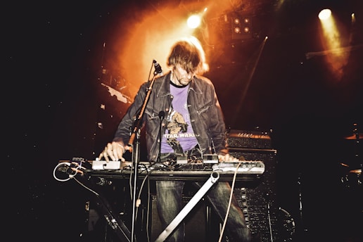 A person is performing on stage with a keyboard and various electronic equipment, surrounded by a warm, orange glowing light. They are wearing a jacket and a graphic t-shirt.