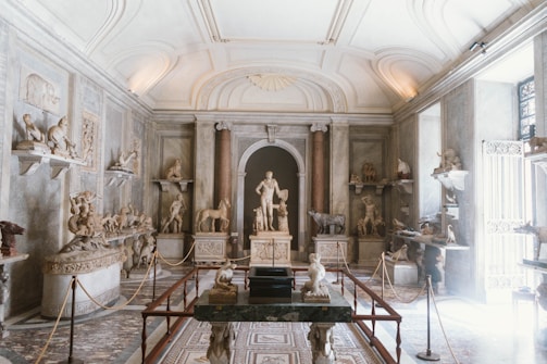 A classical museum gallery filled with various sculptures and artifacts. The room features intricate architectural details, including a high arched ceiling with decorative mouldings and several statues displayed on shelves and pedestals. The light streaming in from the windows creates a serene atmosphere, highlighting the stone textures of the sculptures.