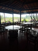 A wooden-furnished indoor area with several round tables and chairs arranged in an open-air setting, offering a scenic view of lush greenery through large windows. The wooden interior and beams create a cozy, rustic atmosphere.