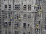 Workers on bamboo scaffolding are repairing or maintaining a building facade. The structure consists of multiple stories, with workers wearing helmets and safety gear.