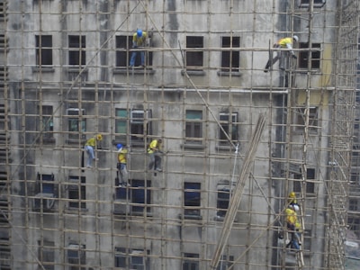 Workers on bamboo scaffolding are repairing or maintaining a building facade. The structure consists of multiple stories, with workers wearing helmets and safety gear.