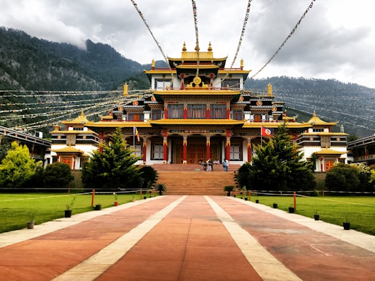 A large, ornate Buddhist monastery with a red and gold color scheme is surrounded by lush greenery and set against a backdrop of misty mountains. Colorful prayer flags are strung above the courtyard, and a group of people are seen on the steps leading to the entrance.