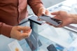 Two hands are exchanging a smartphone over a glass counter. The screen of the phone is cracked, indicating possible damage or need for repair. One person is wearing a brown shirt, and the counter displays other phones and electronic devices.