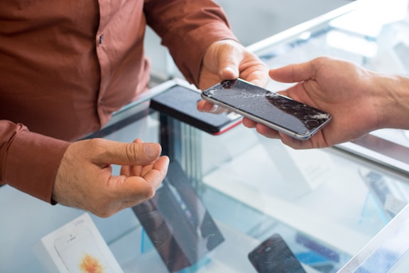 Two hands are exchanging a smartphone over a glass counter. The screen of the phone is cracked, indicating possible damage or need for repair. One person is wearing a brown shirt, and the counter displays other phones and electronic devices.