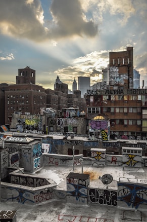 Urban landscape featuring numerous buildings covered in vibrant graffiti. The sky is partly cloudy, with rays of sunlight breaking through the clouds, casting a warm glow over the scene. In the background, distant skyscrapers loom, contrasting with the gritty rooftops in the foreground.