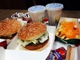 Two burgers with sesame seed buns and lettuce fillings are placed in open boxes. A tray contains fries topped with melted cheese. Two cups with straws are filled with soft drinks. Additional items include two small bowls of ice cream with chocolate drizzle and ketchup packets nearby.