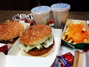 Two burgers with sesame seed buns and lettuce fillings are placed in open boxes. A tray contains fries topped with melted cheese. Two cups with straws are filled with soft drinks. Additional items include two small bowls of ice cream with chocolate drizzle and ketchup packets nearby.