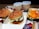 Two burgers with sesame seed buns and lettuce fillings are placed in open boxes. A tray contains fries topped with melted cheese. Two cups with straws are filled with soft drinks. Additional items include two small bowls of ice cream with chocolate drizzle and ketchup packets nearby.