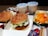 Two burgers with sesame seed buns and lettuce fillings are placed in open boxes. A tray contains fries topped with melted cheese. Two cups with straws are filled with soft drinks. Additional items include two small bowls of ice cream with chocolate drizzle and ketchup packets nearby.