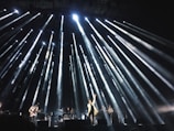 A live concert performance with bright stage lights radiating outward from above, illuminating musicians on stage. The central performer stands at the front with one arm raised, surrounded by band members playing instruments.