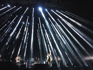 A live concert performance with bright stage lights radiating outward from above, illuminating musicians on stage. The central performer stands at the front with one arm raised, surrounded by band members playing instruments.