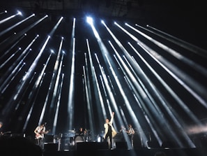 A live concert performance with bright stage lights radiating outward from above, illuminating musicians on stage. The central performer stands at the front with one arm raised, surrounded by band members playing instruments.