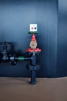 A metal valve with red and green components, mounted on a black pipe against a dark wall. The valve is secured with a chain, and an indicator with a colored diagram is attached above it.