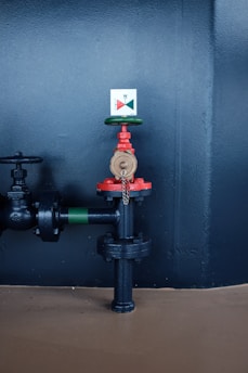 A metal valve with red and green components, mounted on a black pipe against a dark wall. The valve is secured with a chain, and an indicator with a colored diagram is attached above it.