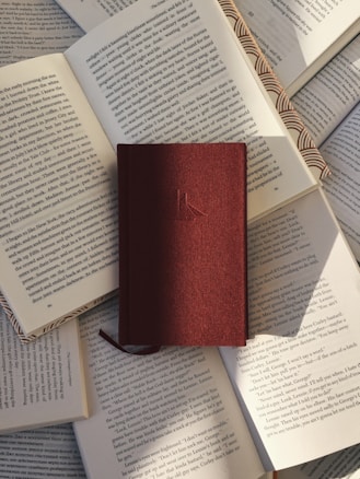 A hardcover book with a maroon cover rests on top of several open books with visible text. The pages of the open books are arranged in a slightly scattered pattern, creating a layered effect. Sunlight casts soft shadows across the books, highlighting the textures of the paper and the cover.