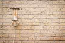 A wall-mounted outdoor light fixture on a textured beige brick wall. The light fixture has a cylindrical glass cover with a visible bulb inside and a metal base plate. A cord is seen running from the fixture downward.