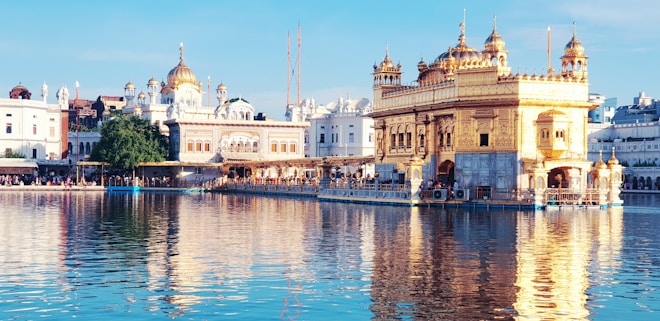A magnificent golden temple is reflected in a tranquil body of water, flanked by other ornate buildings with domes and spires. The morning or late afternoon sunlight bathes the scene in a warm glow, enhancing the ornate details of the architecture. People are visible around the temple, contributing to a serene and sacred atmosphere.