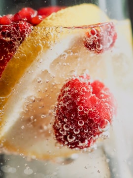 Slices of lemon and raspberries float in a clear liquid with bubbles clinging to the surface. The image captures the refreshing and sparkling nature of the drink, highlighting the vibrant colors and textures of the fruit.