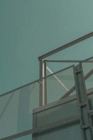 A minimalist architectural scene featuring a mesh metal fence against a pale, muted teal sky. The structure includes angular lines and a metal framework, creating a geometric pattern with the fence's diamond-shaped openings.