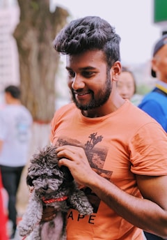 A man wearing an orange t-shirt is smiling while petting a small curly-haired dog. The dog, sporting a red collar, appears comfortable and is held in the man's left arm. The background seems to be an outdoor setting with other people visible but out of focus.