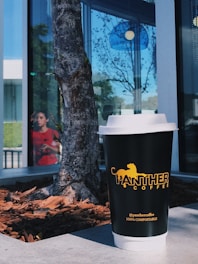 A disposable coffee cup branded with Panther Coffee is placed on a concrete surface beside a tree with mulch at its base. The background shows a modern building with large glass windows reflecting trees and a person wearing a red shirt.