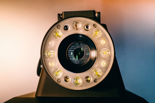 A close-up view of a circular camera lens surrounded by multiple small LED lights. The lens is positioned centrally, with a dark and reflective surface. The LED lights are evenly distributed around the lens, each emitting a soft glow. The background features a gradient of warm and cool tones, creating a contrast with the dark camera housing.