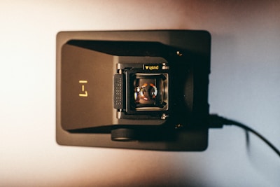 A vintage camera viewed from above, showcasing the lens assembly. The camera rests on a dark surface with a cable connected to its side. The branding 'Impossible' is visible, along with a button labeled 'push.'