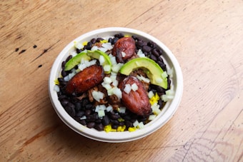 A bowl filled with black beans, sliced avocado, fried plantains, chopped onions, and what appears to be yellow rice or possibly shredded cheese. The ingredients are arranged in a visually appealing manner, with the colors contrasting against each other. The bowl is set on a wooden surface, which adds a rustic element to the presentation.